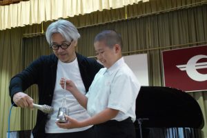 男子生徒が手にもった道具で音を鳴らし、坂本龍一氏がマイクを道具にあて音をひろっている生徒との交流の様子を写した写真