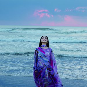 ピンク色に染まる雲、穏やかに押し寄せる波をバックに砂浜に立つ紫系のドレスを着た小田 加奈子さんの写真