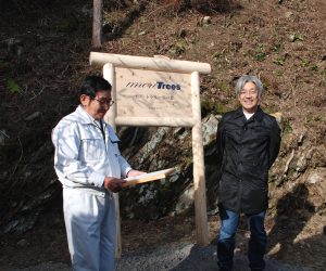 more treesと書かれた看板の横に立つ関係者の男性と笑顔の坂本龍一氏の写真