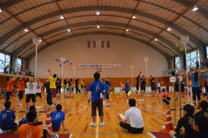 「九州玉入れ選手権大会inもろつか」と書かれた幕が掲げられた体育館で6つのグループがそれぞれのカゴをめがけて玉を投げている写真