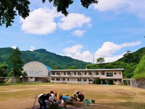 奥に体育館と校舎が見える校庭で、生徒達が地面にしゃがみ込み活動を行っている様子の写真