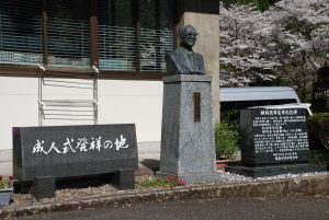 横長で黒色の石碑に白字で「成人式発祥の地」と書かれている石碑と、その右隣りに藤井長次郎氏の胸像が建っている写真