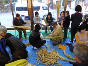 ブルーシートが広げられた場所で年配の男性がしめ縄を作っている様子を、傍でじっと見ている子供たちの写真