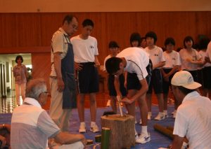 大人の男性達が見守る中、男子中学生が丸太の上で棒のような物を裂いている様子の写真