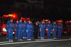 夜に赤色の回転灯を付けた消防車が横に並んで駐車されており、青い消防服を着た消防団の方々が整列し敬礼している写真