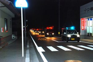夜に青色の回転灯を付けた軽自動車を先頭に後ろには赤色の回転灯を付けた車が道路の後方まで続いて走行しているパレードの様子の写真