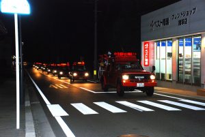 夜の道路で、赤色の回転灯を付けた消防車が何台も後ろの方まで続いているパレードの様子の写真