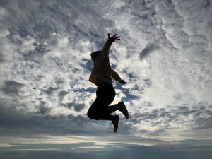 空には白い筋状の雲や積雲があり、両手を広げ大きくジャンプしている人が黒い描写で写っている写真