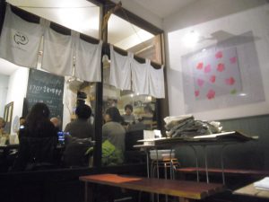 白い正方形の形が6枚並びになっている暖簾が窓ガラスに掛かっており、店内にはお客さんが席につき賑わっている様子の写真