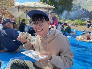 参加者の男性が串にさして焼いた焼き魚を食べようとして、満面の笑顔で写っている写真