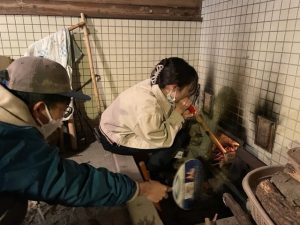 男性がうちわであおぎ、女性が火吹き棒から息を吹きかけ火起こしをしている写真