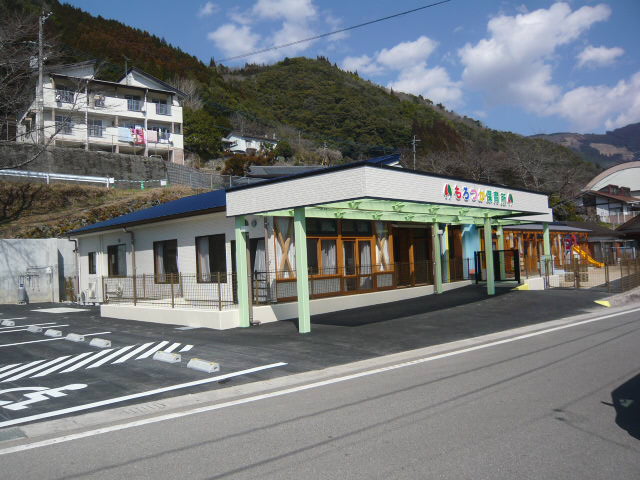 左側に駐車場があり、白い壁青い屋根の園舎で、道路に面した側にせり出した庇のような部分に「もろつか保育所」とカラフルな色で書かれている諸塚保育所の外観写真