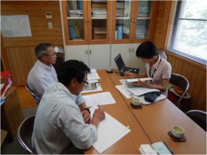 向かい合わせに並べた2つの長机の席に3名の方が座り、書類を広げながら話し合いをしている写真
