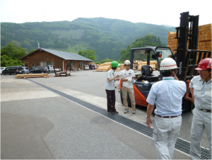 奥に平屋建ての建物、木材などが置かれている広い敷地のフォークリフトの横で手に書類を持った職員の方が、加工センターの職員へ聞き取りを行っている写真