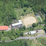 山に囲まれた自然豊かな場所に運動場のような広場と横にL字型の建物、赤い屋根の建物が見える航空写真