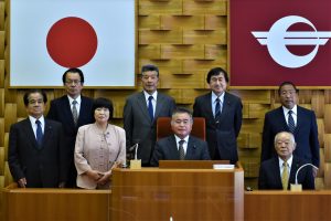 日本の国旗と諸塚村の町旗が掲示されている議会の議長席前に関係者の方々8名が集まり写っている記念写真